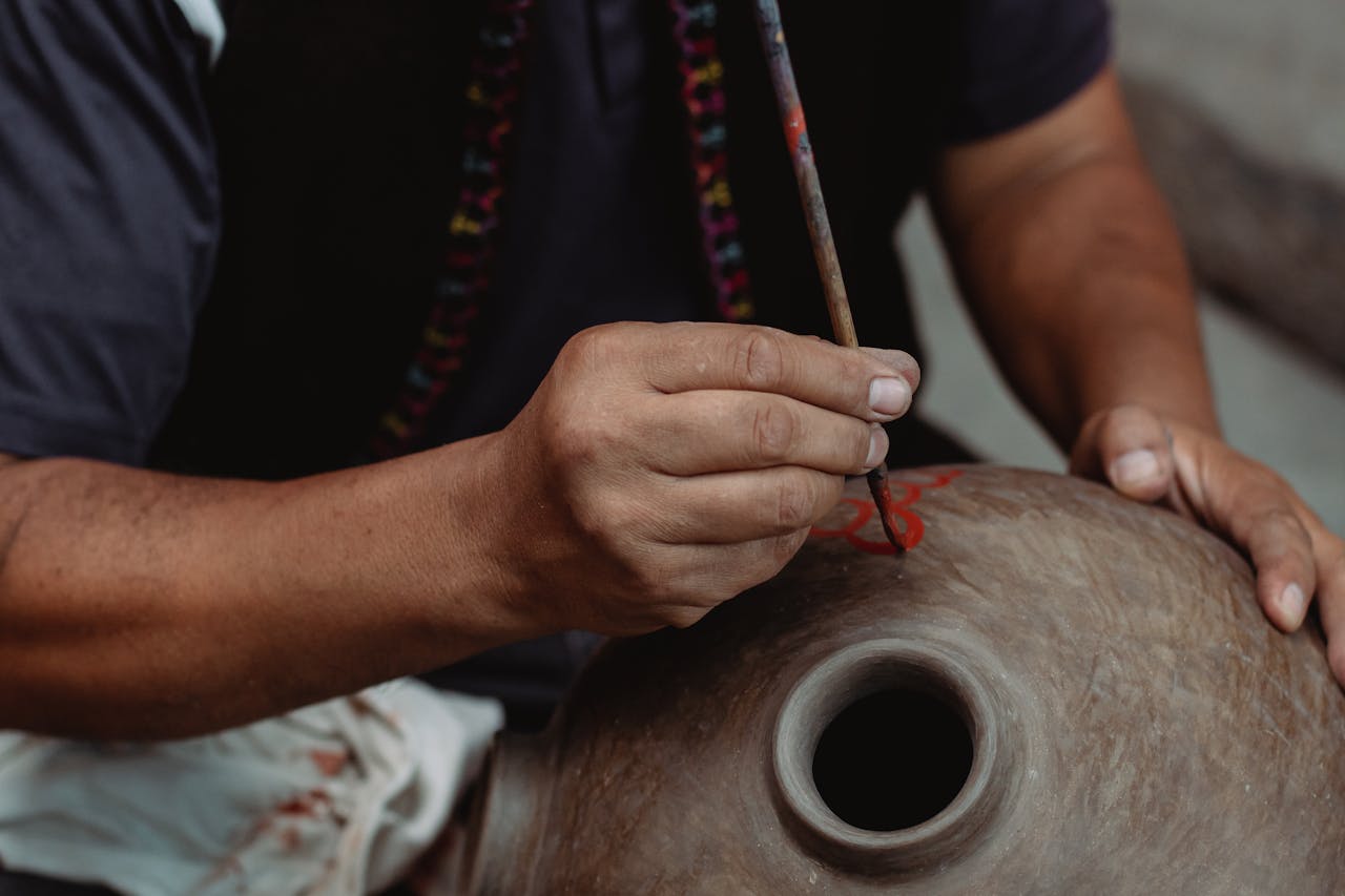 A close-up of an artisan's hands painting a handmade clay jar using a paintbrush.